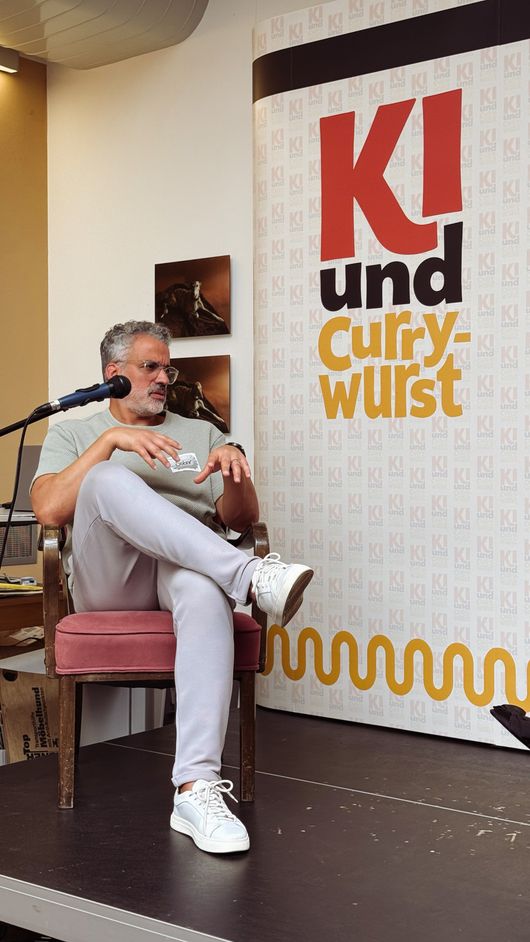 Moderator Saber Romdhani auf der Bühne vor dem KI-und-Currywurst-Banner