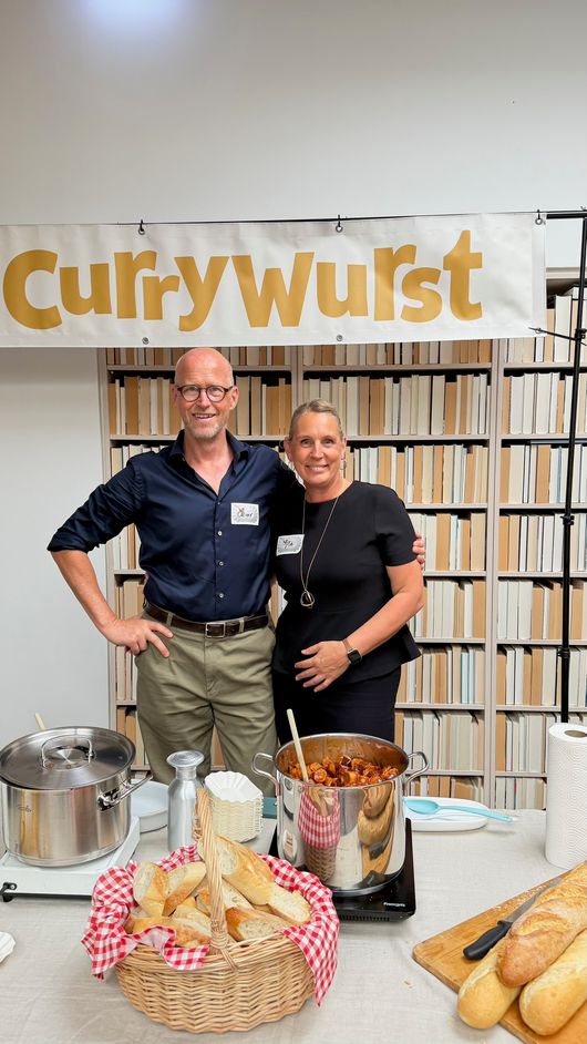 Zwei Gastgeber an der Currywurst-Station vor dem Event-Banner